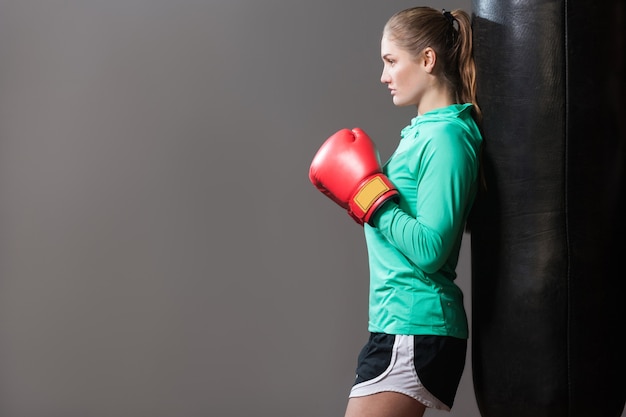 Vista laterale del profilo di una giovane donna seria atleta con i capelli raccolti in maniche lunghe verdi e pantaloncini neri appoggiati al sacco da boxe in guanti rossi da boxe. Al coperto, isolato su sfondo grigio scuro