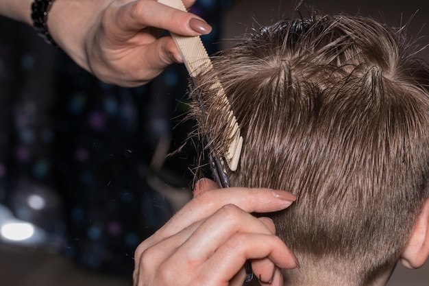 Vista laterale del ragazzino carino che si fa tagliare i capelli dal parrucchiere dal barbiere usando il pettine e le forbici da toelettatura. Primo piano, il concetto di moda e bellezza.
