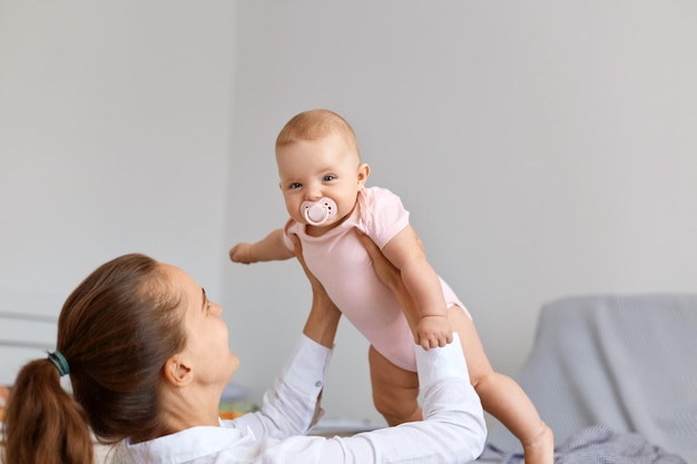 Vista laterale del ritratto di donna felice positiva con i capelli scuri donna che gioca con la figlia neonata a casa, vomita un bambino in aria, trascorre del tempo a casa.