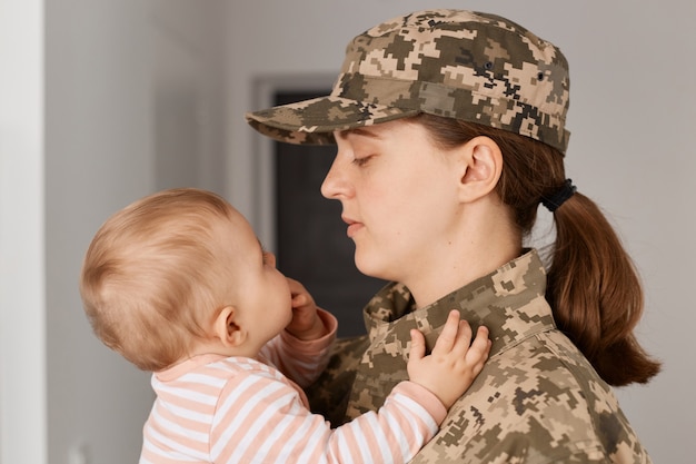 Vista laterale del ritratto di un'adorabile giovane donna soldato adulta che indossa l'uniforme mimetica, torna a casa e incontra la sua piccola figlia, guardando la figlia, non posso credere al loro incontro.