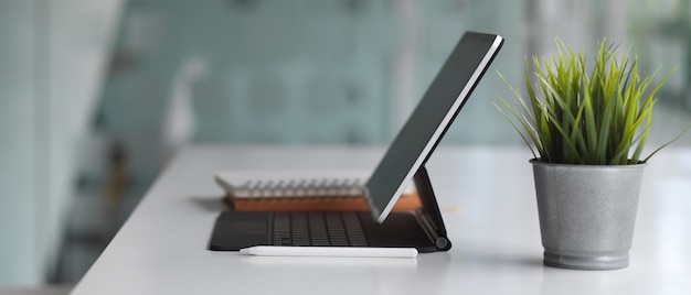 Vista laterale del tavolo bianco con tavoletta digitale, tastiera, taccuino e vaso per piante nello spazio di lavoro co