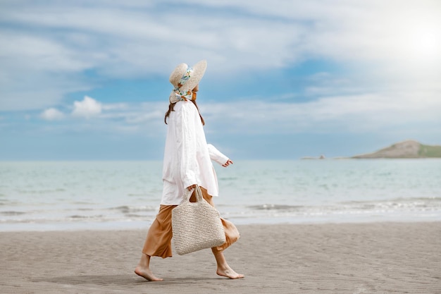 Vista laterale del viaggiatore allegro della giovane donna con il cappello di paglia che cammina sulla spiaggia durante la vacanza