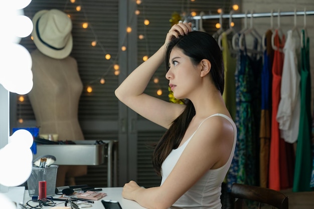 vista laterale dell'affascinante splendida artista coreana di successo che corregge i suoi capelli seduti al tavolo da toeletta guardando all'interno dello specchio riflesso nello spogliatoio. host di signora asiatica nel trucco della canotta dietro le quinte