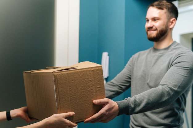 Vista laterale dell'amichevole corriere barbuto che consegna pacchi in scatola di cartone al cliente femminile a due passi da casa