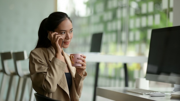 Vista laterale dell'imprenditore femminile che parla al telefono mentre fa una pausa nella stanza dell'ufficio della parete di vetro