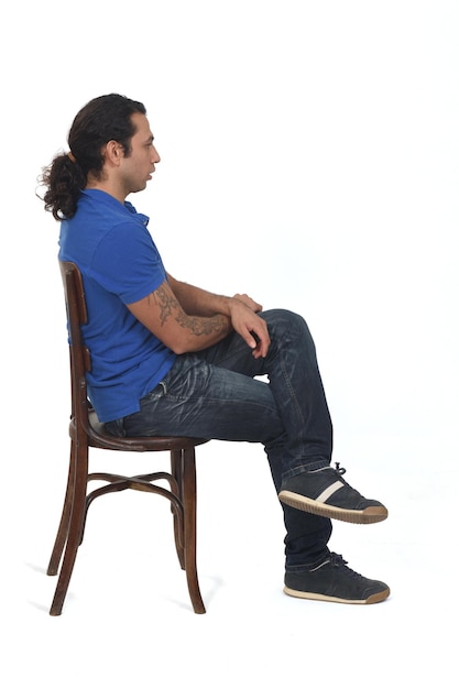 Vista laterale dell'uomo con coda di cavallo e abbigliamento casual seduto su una sedia con le gambe incrociate su sfondo bianco