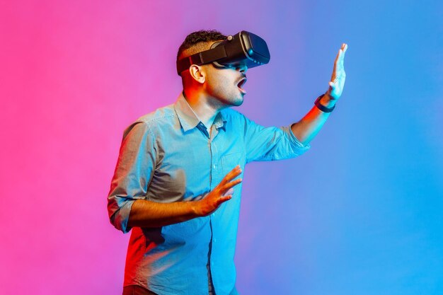 Vista laterale dell'uomo con gli occhiali per realtà virtuale sulla testa che gioca al simulatore di gioco in piedi mani tese che toccano l'oggetto nel cyberspazio Studio indoor girato isolato su sfondo colorato di luce al neon