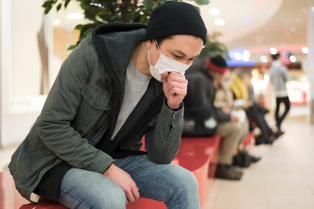 Vista laterale dell'uomo con la maschera medica che tossisce mentre al centro commerciale