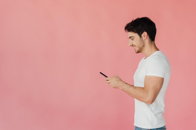 Vista laterale dell'uomo muscoloso sorridente in maglietta bianca utilizzando smartphone isolato su rosa