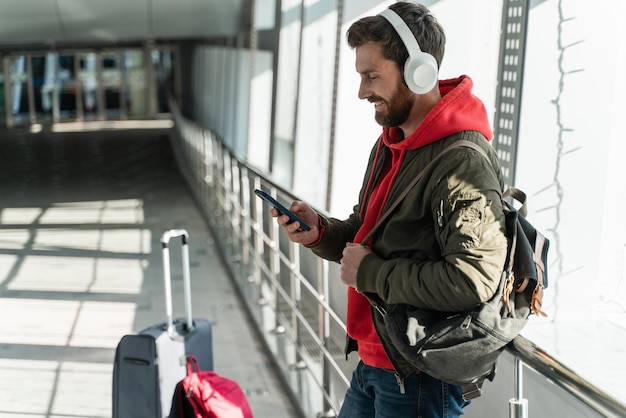 Vista laterale dell'uomo turistico che indossa abiti casual ascoltando musica tramite le cuffie mentre si appoggia alla finestra e aspetta il suo volo Foto d'archivio