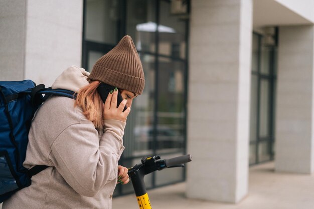 Vista laterale della consegna di cibo da corriere femminile in abiti caldi che chiama al cliente sul telefono cellulare in piedi in strada con scooter elettrico