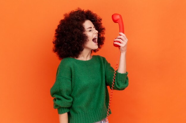 Vista laterale della donna arrabbiata con l'acconciatura afro che indossa un maglione verde in stile casual che urla mentre parla con il cliente nel call center emozioni negative Studio interno girato isolato su sfondo arancione