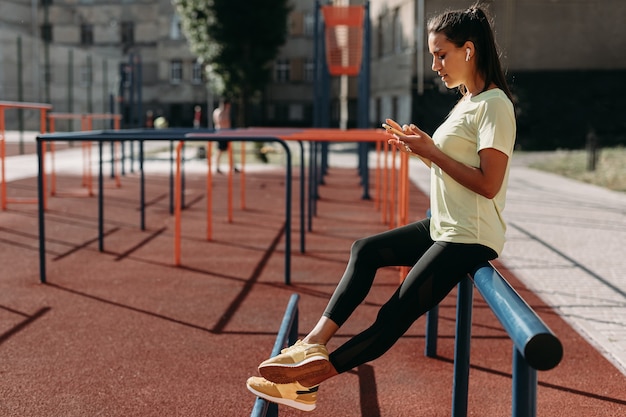 Vista laterale della donna atletica stanca con i capelli scuri utilizza lo smartphone mentre ci si rilassa al campo sportivo.