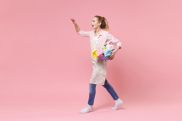 Vista laterale della donna casalinga nel grembiule con bacinella con bottiglie di detersivo che lavano i detergenti facendo i lavori domestici isolati su sfondo rosa. Concetto di pulizia. Salutare con la mano mentre nota qualcuno.