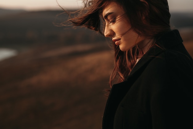 Vista laterale della donna delicata in cappotto e con i capelli volanti in piedi nella natura al tramonto in autunno e guardando in basso nella contemplazione