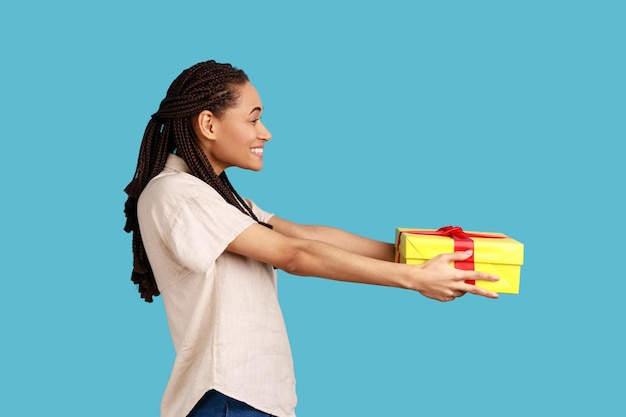 Vista laterale della donna felice con i dreadlocks neri che danno una confezione regalo avvolta e sorride davanti