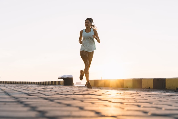 Vista laterale della donna fitness in esecuzione su una strada in riva al mare vicino al porto marittimo. Allenamento sportivo sul lungomare al tramonto.