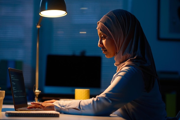 Vista laterale della donna musulmana che lavora online sul computer portatile in ufficio durante la scadenza