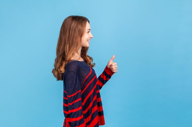 Vista laterale della donna sorridente che indossa un maglione a righe in stile casual che distoglie lo sguardo mostrando il pollice in su esprimendo approvazione feedback eccellente Colpo in studio indoor isolato su sfondo blu