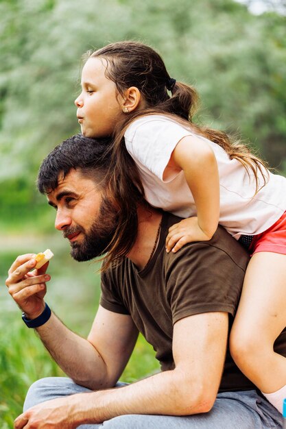 Vista laterale della famiglia sorridente seduta sul picnic nella foresta del parco intorno agli alberi Piccola figlia seduta sulla schiena dei padri