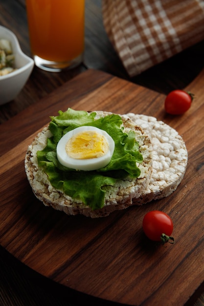 Vista laterale della fetta di uovo con lattuga su pane croccante e pomodori sul tagliere con succo d'arancia su sfondo di legno