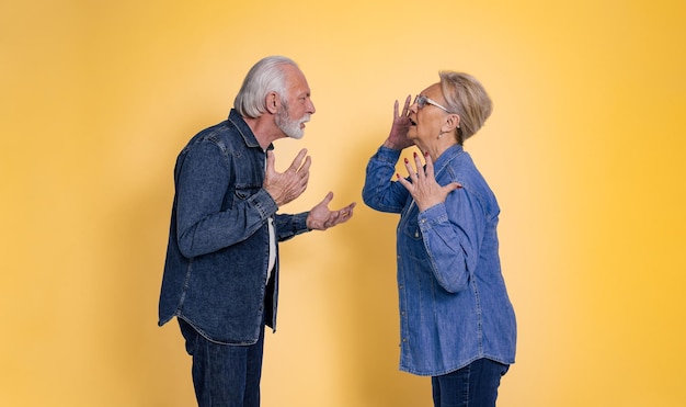 Vista laterale della furiosa coppia anziana vestita di jeans blu che litigano e urlano l'un l'altro mentre si trovano faccia a faccia isolati su sfondo giallo