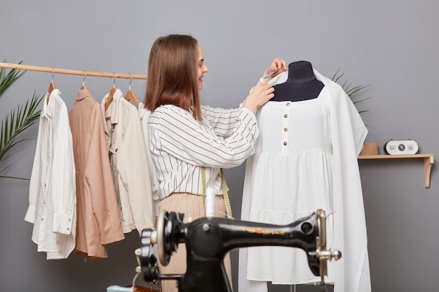 Vista laterale della gioiosa sarta donna professionista che mette il perno in tessuto appeso al manichino mentre si lavora in un moderno laboratorio creando una nuova collezione di abiti da cucire