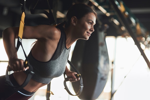 Vista laterale della giovane bella donna atletica che indossa abiti sportivi che si esercita con cinghie trx fitness in palestra industriale, sta allenando le braccia e sorridendo. Sport, allenamento, stile di vita sano