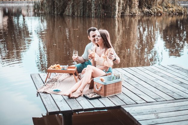 Vista laterale della giovane coppia felice su un picnic romantico vicino al fiume o al lago, donna e uomo che bevono vino all'aperto insieme, persone che si divertono in vacanza, amore