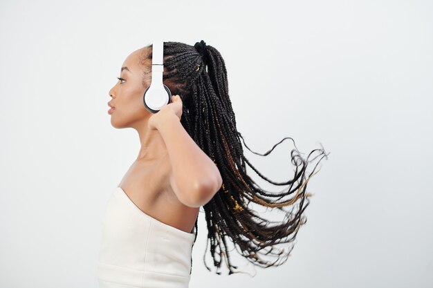 Vista laterale della giovane donna afro-americana in abito bustier bianco ascoltando musica in cuffia