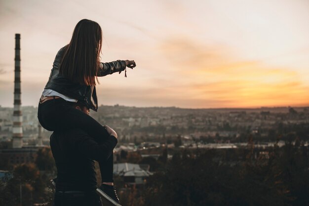 Vista laterale della giovane donna anonima che si siede sulle spalle del ragazzo e che punta al tramonto incredibile sulla città moderna