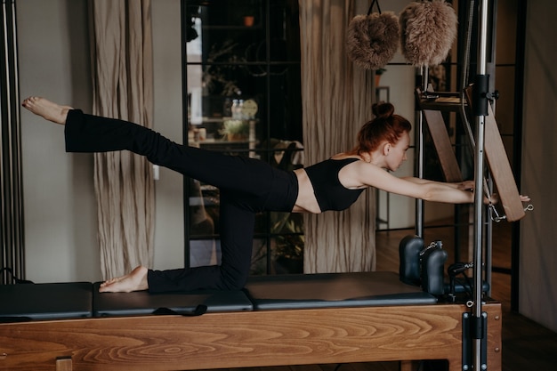 Vista laterale della giovane donna atletica che allunga le gambe su una macchina per pilates o un riformatore Cadillac, indossando top e pantaloni neri mentre si allena in palestra o in palestra, preparando i muscoli per l'allenamento