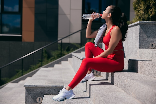 Vista laterale della giovane donna atletica che indossa top rosso e leggins e scarpe da ginnastica bianche sedute sulle scale. Ragazza che si prende una pausa e beve acqua dalla bottiglia di plastica.