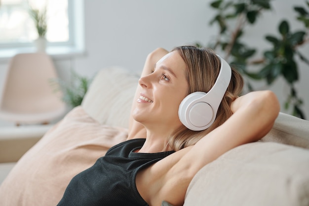 Vista laterale della giovane donna bionda sorridente in cuffia che si gode la musica mentre è sdraiata sul retro del divano in ambiente domestico e si riposa