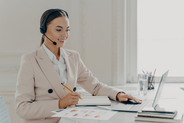 Vista laterale della giovane donna d'affari sorridente che indossa prendendo parte a una conferenza web, guardando lo schermo del laptop, operatore di call center che consulta il cliente mentre è seduto dietro la scrivania in un ufficio moderno