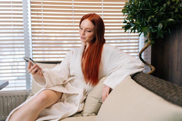 Vista laterale della giovane donna felice che indossa un accappatoio bianco utilizzando il telefono cellulare che guarda sorridente allo schermo del dispositivo seduto vicino alla finestra sul comodo divano del centro benessere