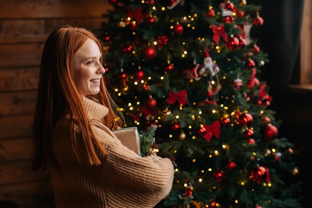 Vista laterale della giovane donna felice che tiene bella confezione regalo di Natale sullo sfondo dell'albero di Natale