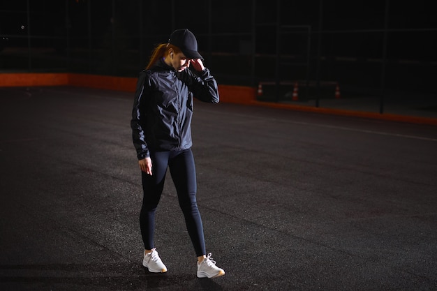 Vista laterale della giovane donna in abbigliamento sportivo e berretto che cammina riposandosi all'aperto facendo una pausa a ni...