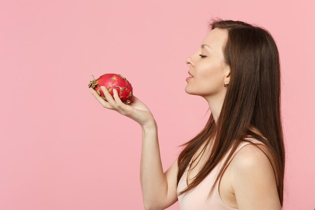 Vista laterale della giovane donna in abiti leggeri che tiene frutta fresca matura del drago pitahaya isolata su sfondo rosa pastello in studio. Lo stile di vita vivido delle persone si rilassa il concetto di vacanza. Mock up spazio di copia.