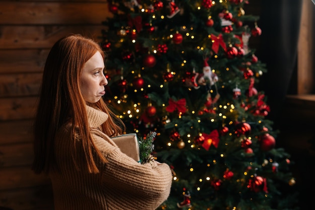 Vista laterale della giovane donna insoddisfatta che tiene il contenitore di regalo di Natale sullo sfondo dell'albero di Natale