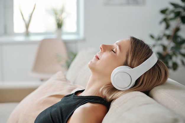 Vista laterale della giovane donna rilassata in cuffia sdraiata sul retro del divano e ascoltando musica rilassante mentre si gode il riposo a casa