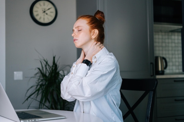 Vista laterale della giovane donna rossa sofferente con gli occhi chiusi che hanno dolore al collo durante il lavoro al computer portatile mentre si è seduti alla scrivania nel posto di lavoro a casa. Concetto di lavoro a distanza da casa.