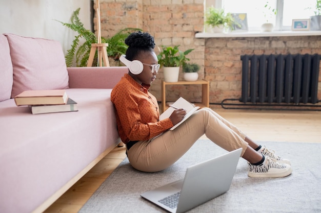 Vista laterale della giovane studentessa africana in cuffia seduta sul pavimento vicino al divano con il computer portatile davanti