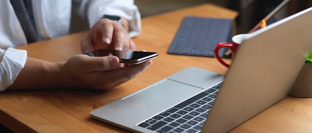 Vista laterale della mano maschio utilizza lo smartphone mentre è seduto al tavolo da lavoro con laptop e forniture per ufficio