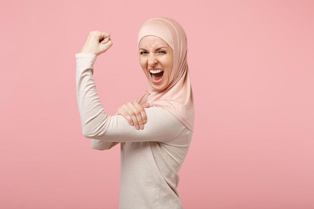 Vista laterale della pazza donna musulmana araba in abiti leggeri hijab in posa isolata su sfondo rosa. Concetto di stile di vita religioso dell'Islam della gente. Mock up spazio di copia. Urlando mostrando bicipiti, muscoli.