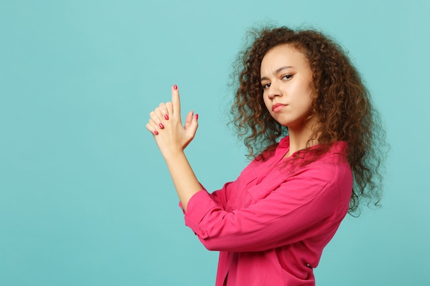 Vista laterale della ragazza africana sicura in abiti casual rosa che si tengono per mano come pistola isolata sul fondo della parete blu turchese in studio. Persone sincere emozioni, concetto di stile di vita. Mock up copia spazio.