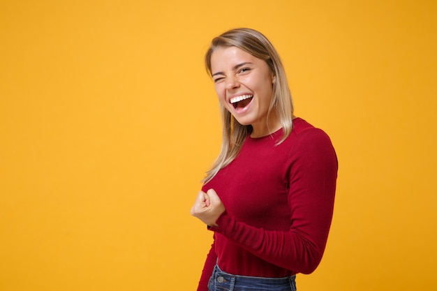 Vista laterale della ragazza felice giovane donna bionda in abiti casual posa isolato su sfondo giallo arancione in studio. Concetto di stile di vita delle persone. Simula lo spazio della copia. Pugno serrato come vincitore, sbattendo le palpebre.