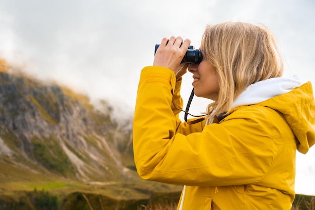 Vista laterale della ragazza in viaggio che guarda attraverso il binocolo in montagne coperte da nuvole in una giornata di sole