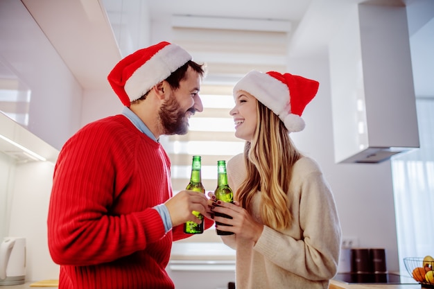 Vista laterale delle coppie adorabili con i cappelli di Santa sulle teste che stanno nella cucina, parlando e bevendo birra
