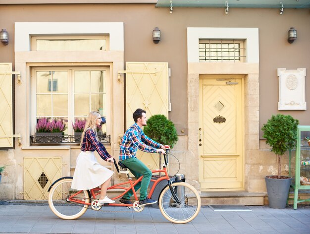 Vista laterale delle coppie turistiche attive, dell'uomo barbuto e della donna bionda che ciclano bici in tandem lungo il marciapiede pavimentato vuoto il giorno di estate soleggiato luminoso dalle case residenziali della città.